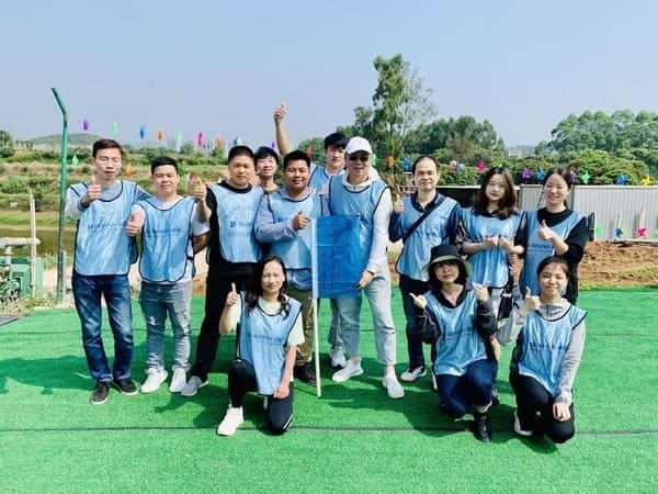 A group of company employees wearing blue vests posing for a group photo during an outdoor team-building activity, holding a blue flag with green fields and blue sky in the background.